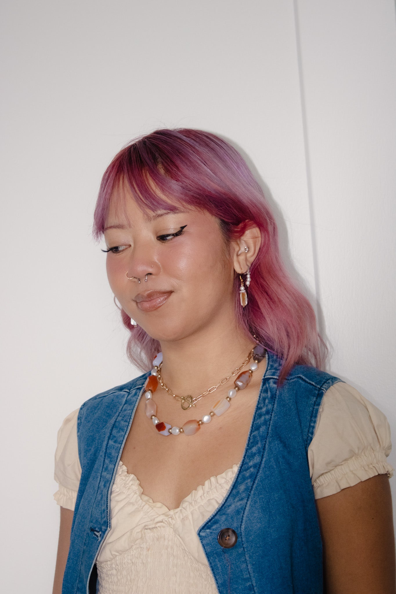 Woman with pink hair wearing a blue vest and beige top against a white background