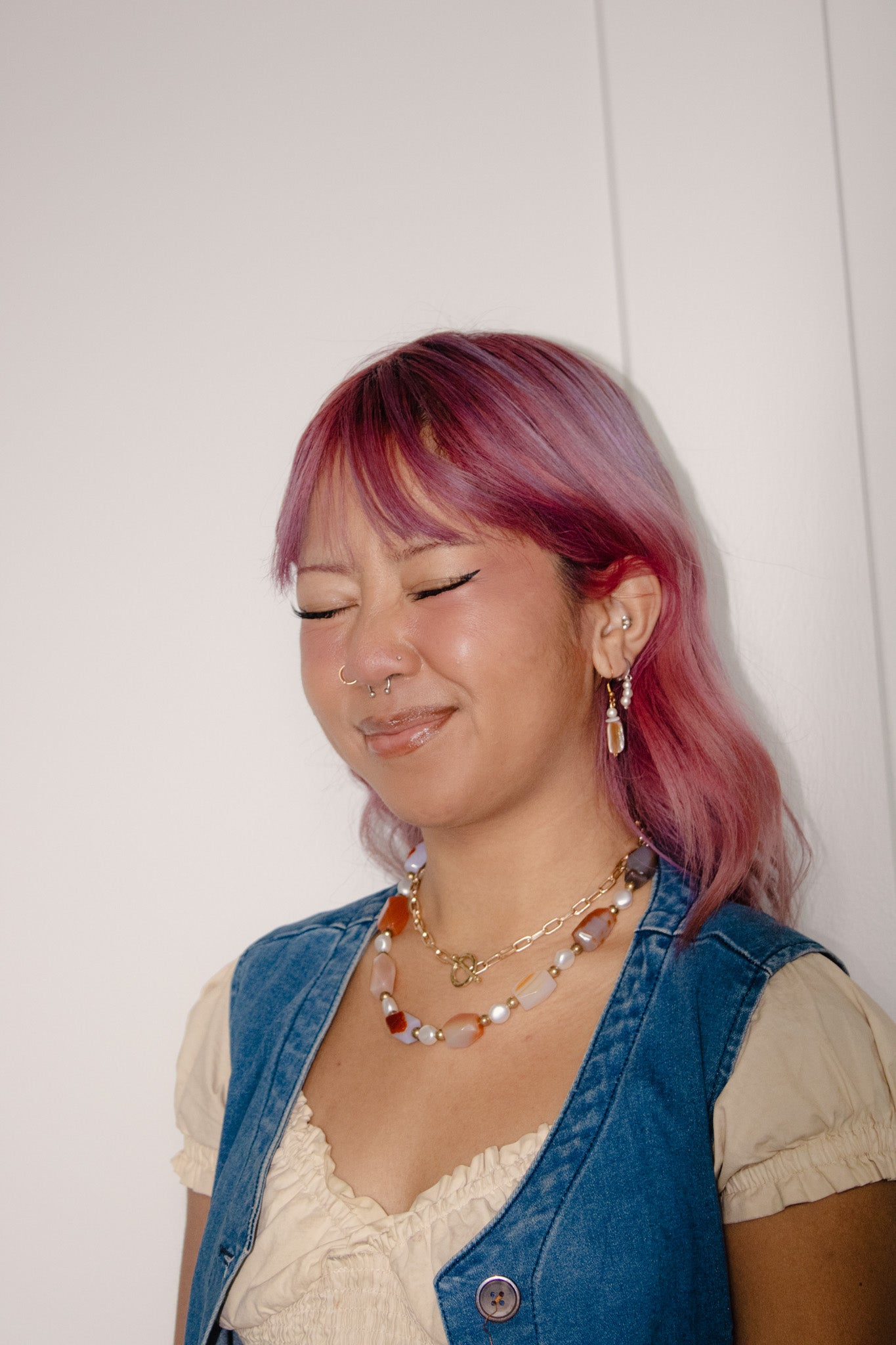 Person with pink hair wearing a denim vest and layered necklaces against a white background