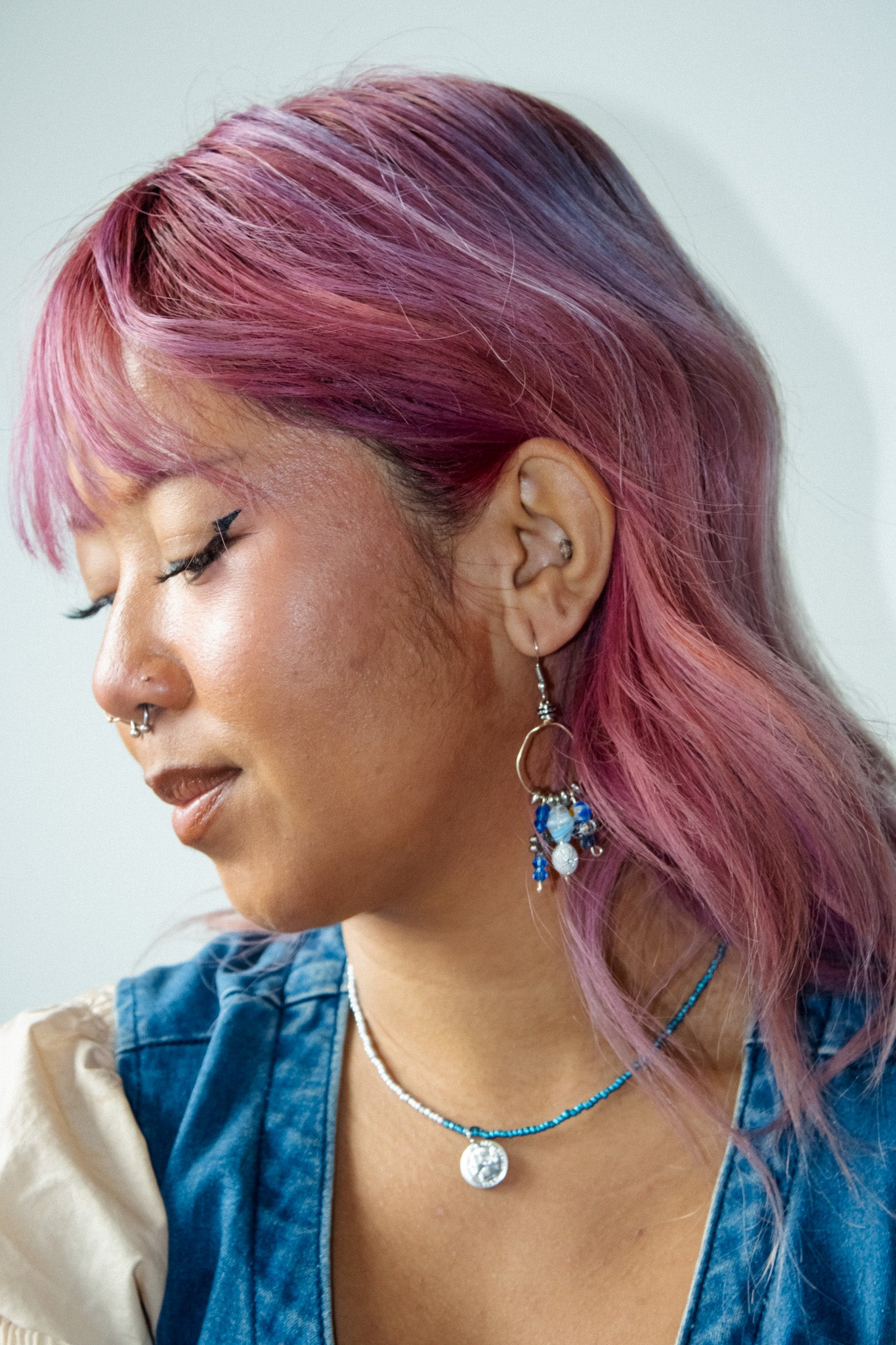 Person with pink hair wearing a blue jacket and colorful earrings against a white background