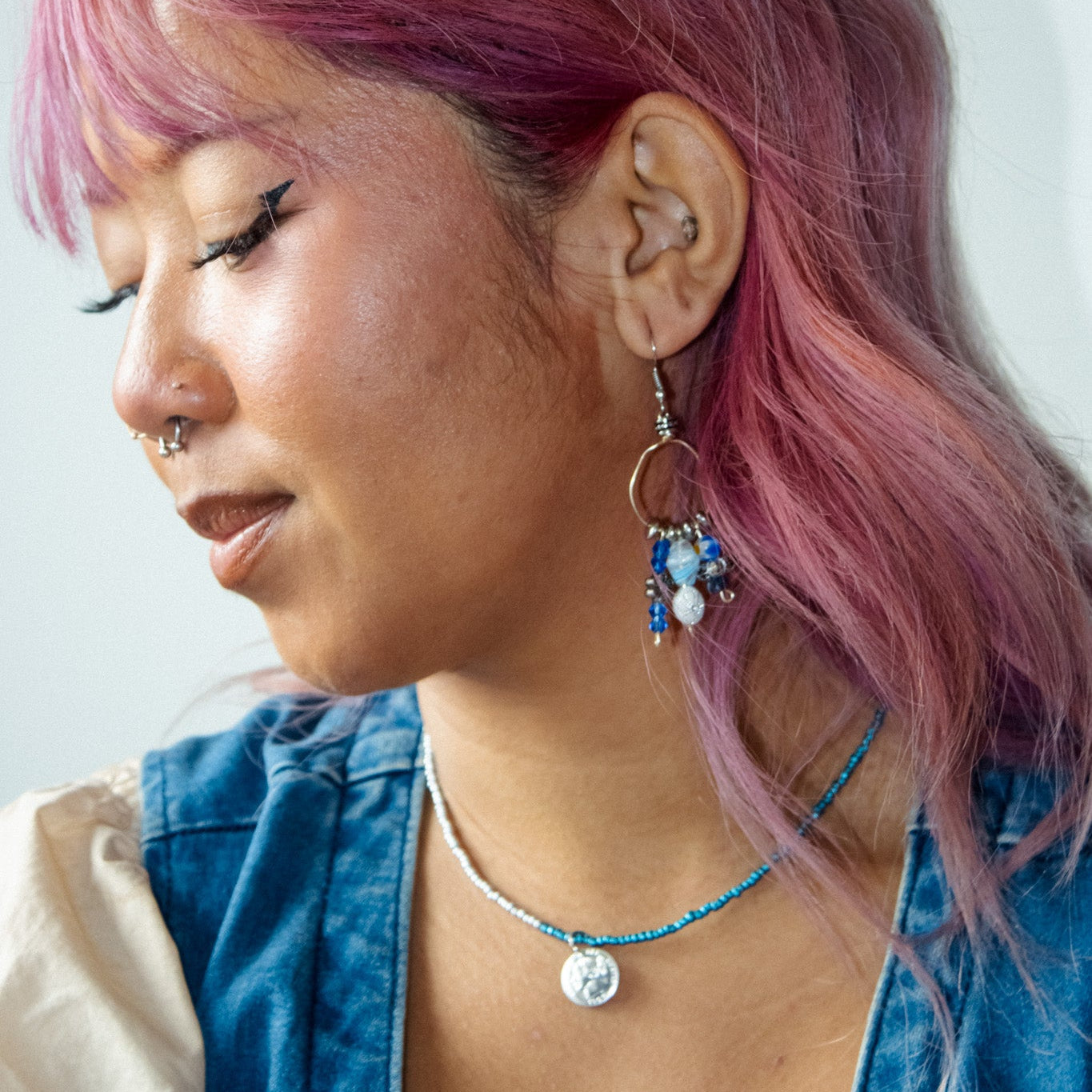 Person with pink hair wearing a blue jacket and colorful earrings against a white background