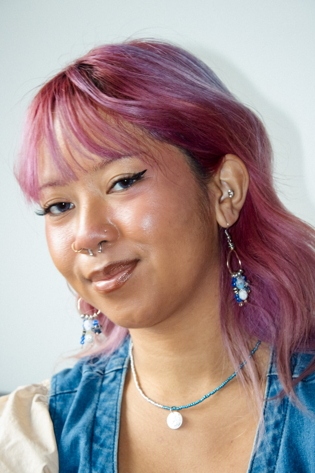 Woman with pink hair wearing a blue denim jacket and colorful earrings.