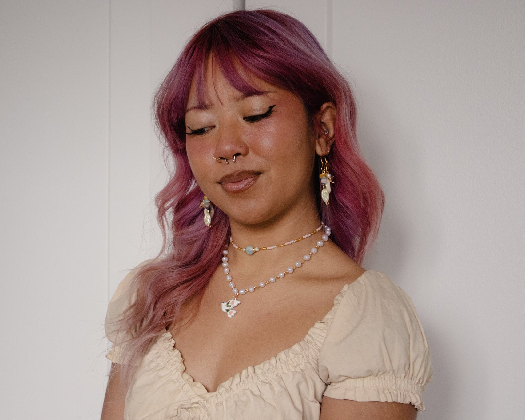 Woman with pink hair wearing a beige top and multiple necklaces against a white background