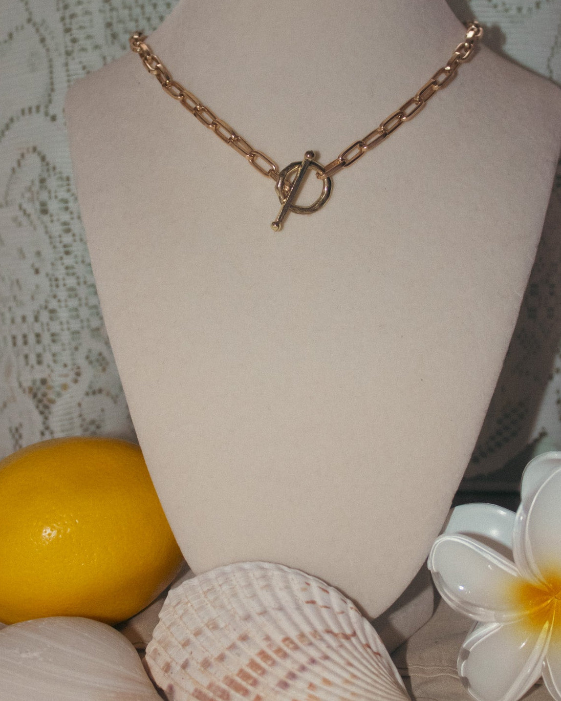 Gold chain necklace on a mannequin bust with shells and a lemon on a textured surface.