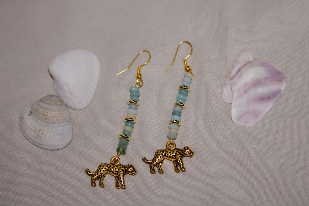 Gold earrings with beaded accents and leopard charms on a gray surface with shells.