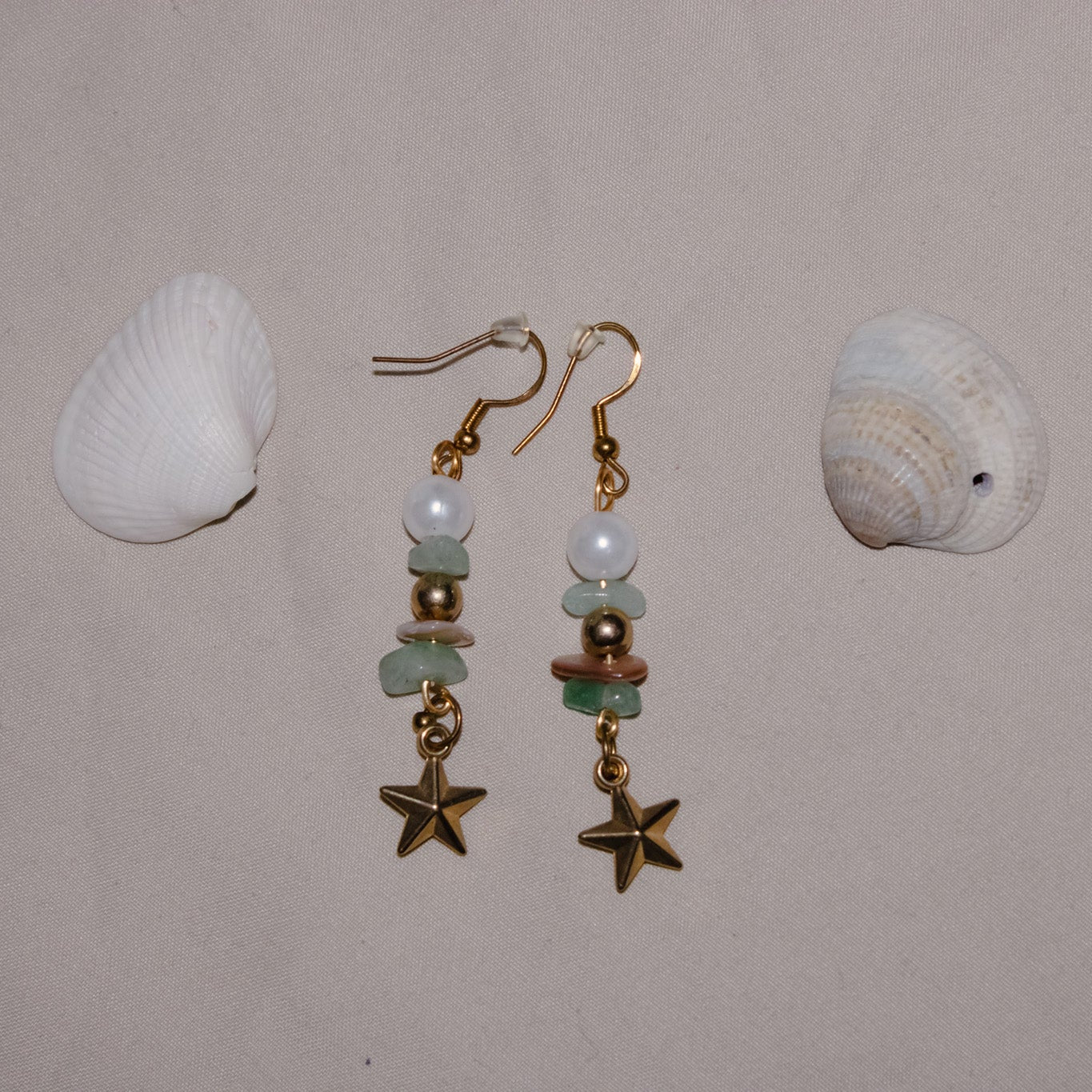 Pair of gold starfish earrings with green beads on a beige background with two shells.