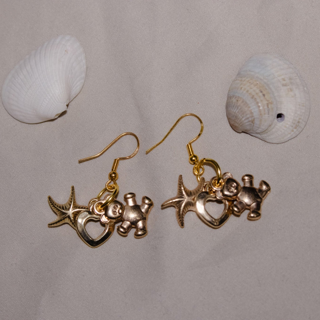 Gold earrings with starfish and heart charms on a gray background with shells.