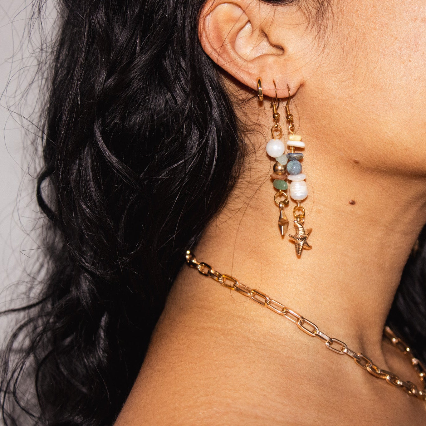 Close-up of a person wearing gold earrings and a chain necklace with a neutral background